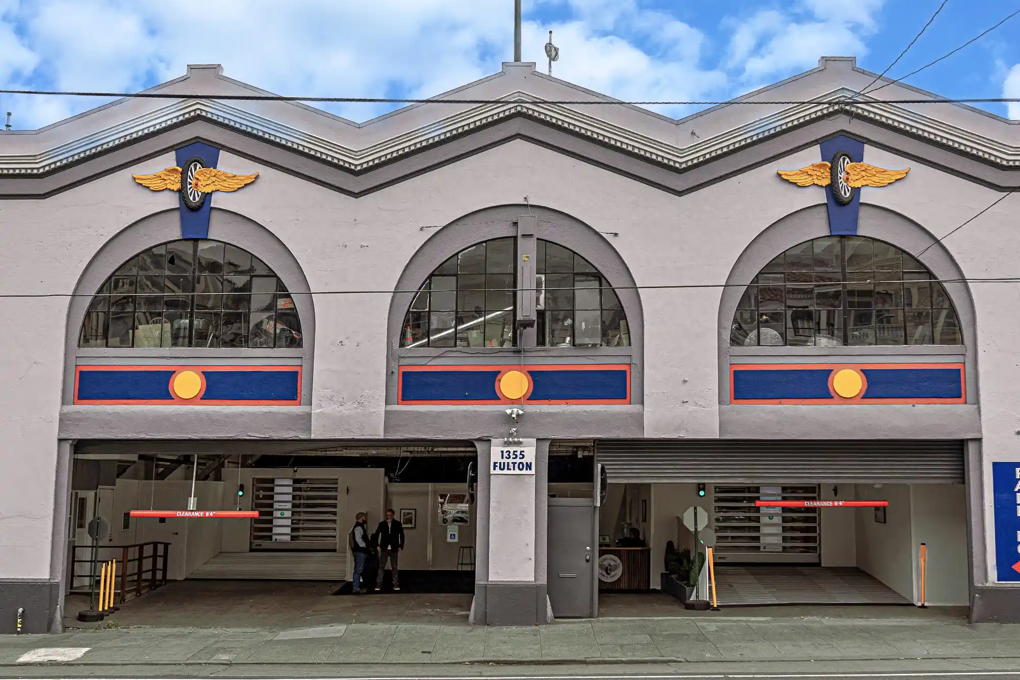 A two-story gray building with large arched windows, blue and gold winged emblems, and an open garage entrance at street level. Three people stand just inside the entrance under a sign reading “1555 Fulton.”.