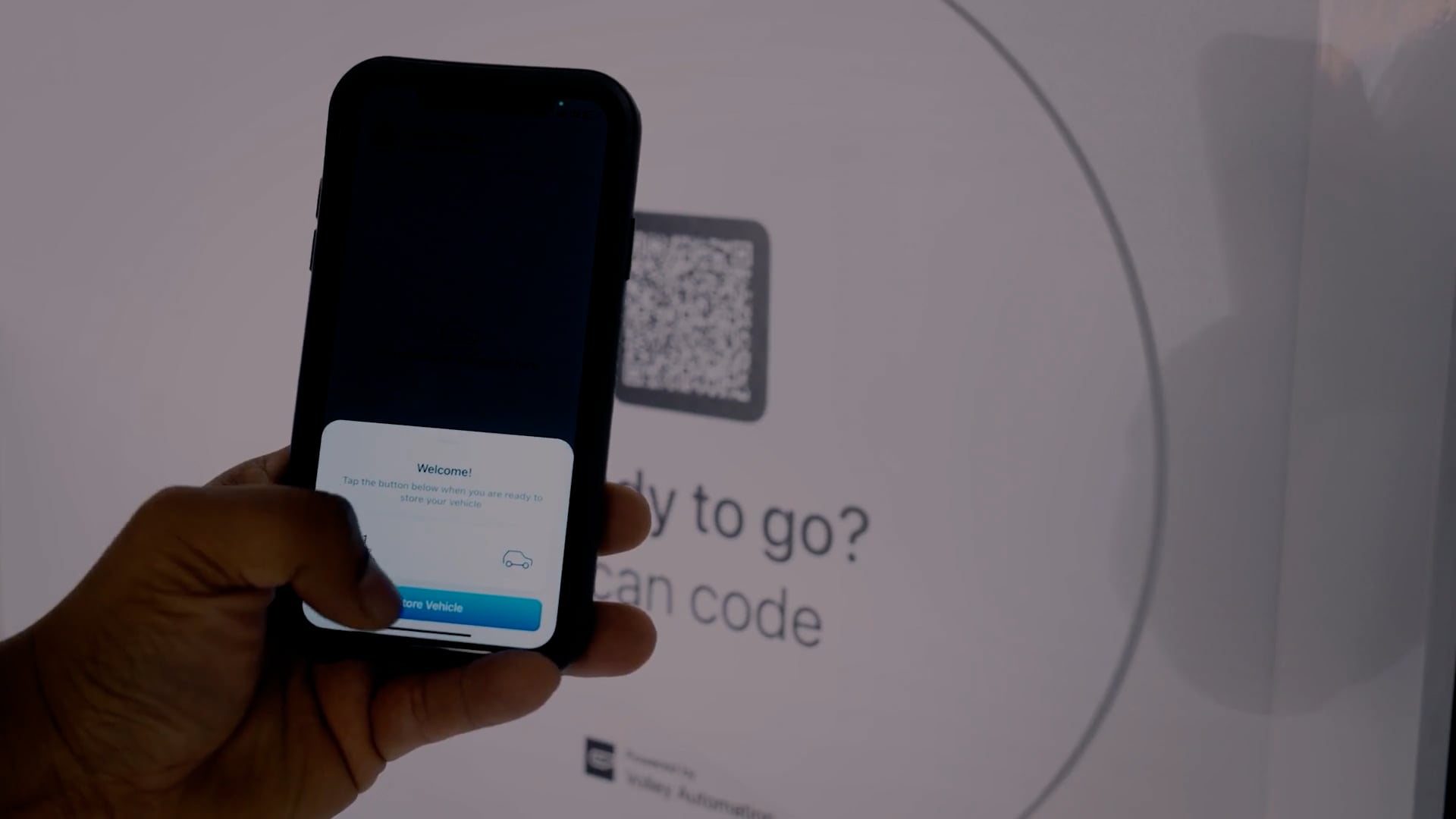 A person's hand holding a smartphone with a notification on the screen, near a digital screen displaying a qr code. The environment suggests an interactive or digital transaction setup.