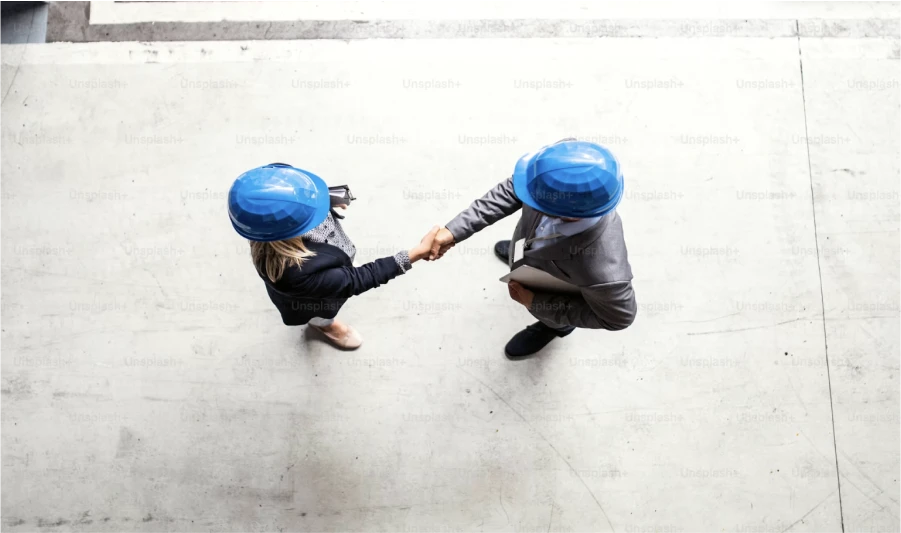 Two professionals wearing blue hard hats shaking hands while standing, viewed from a top-down perspective on a gray concrete background.
