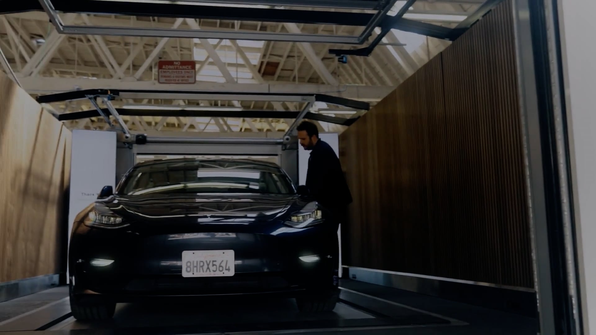 A man in a dark suit stands beside a dark colored car in Volley Automation parking bay with overhead guide rails, in a garage-like setting.