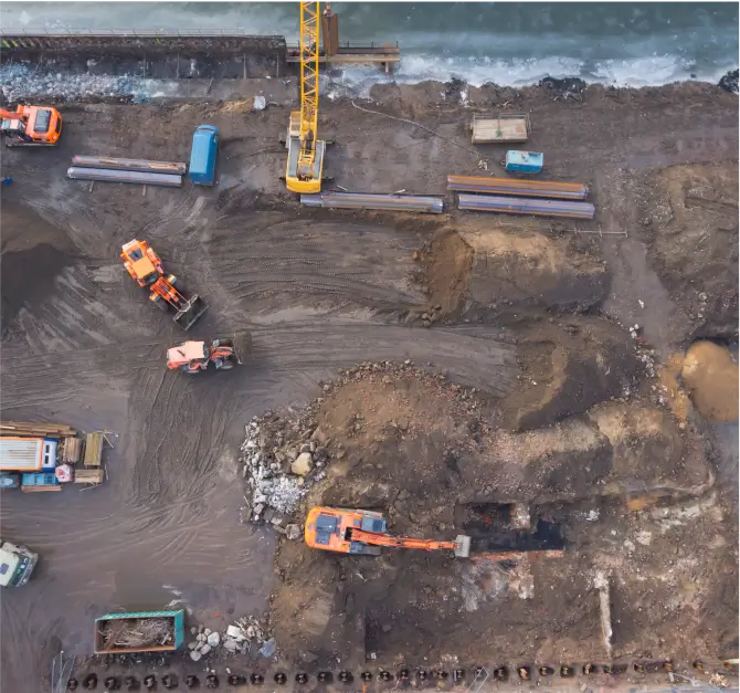 Aerial view of a construction site with various heavy machinery, including excavators and trucks, operating on muddy terrain near a body of water.