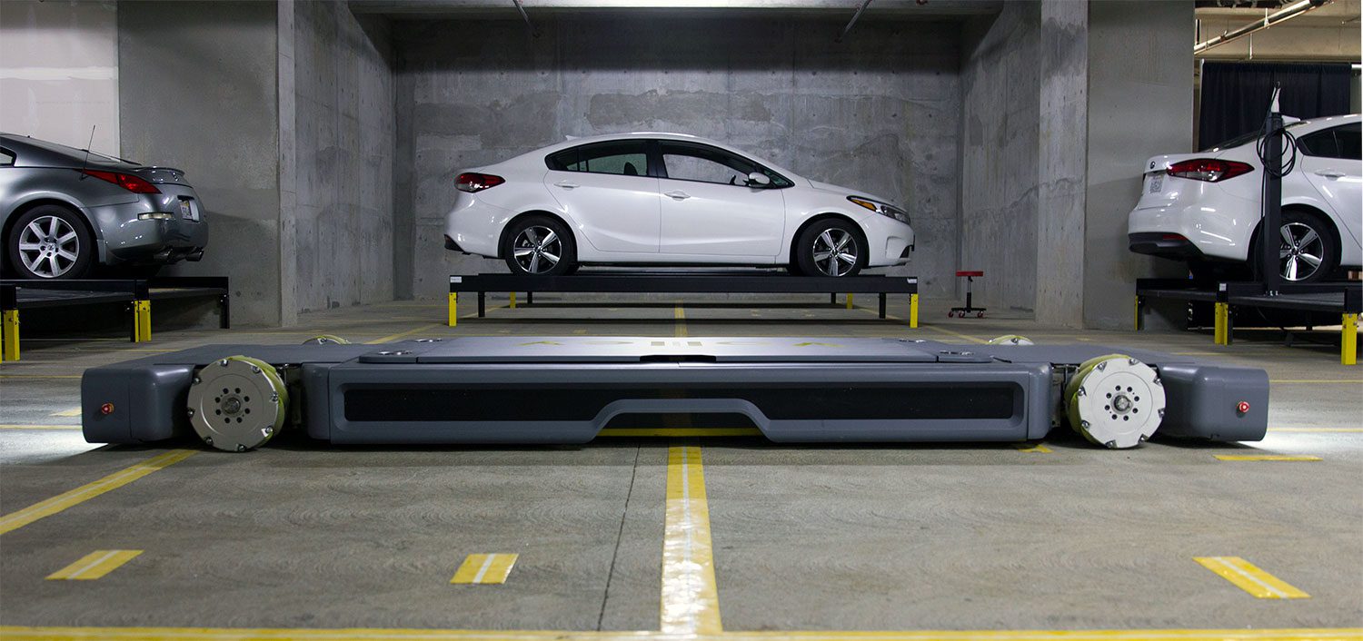 An image of a white car parked in an indoor automated parking lot, facing a large, mobile robot designed for moving parked vehicles. Other cars are visible in the garage.