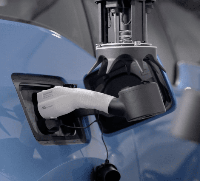 An electric vehicle charging station connector plugged into the charging port of a blue car, demonstrating an automated robotic recharging process with visible cables and port.