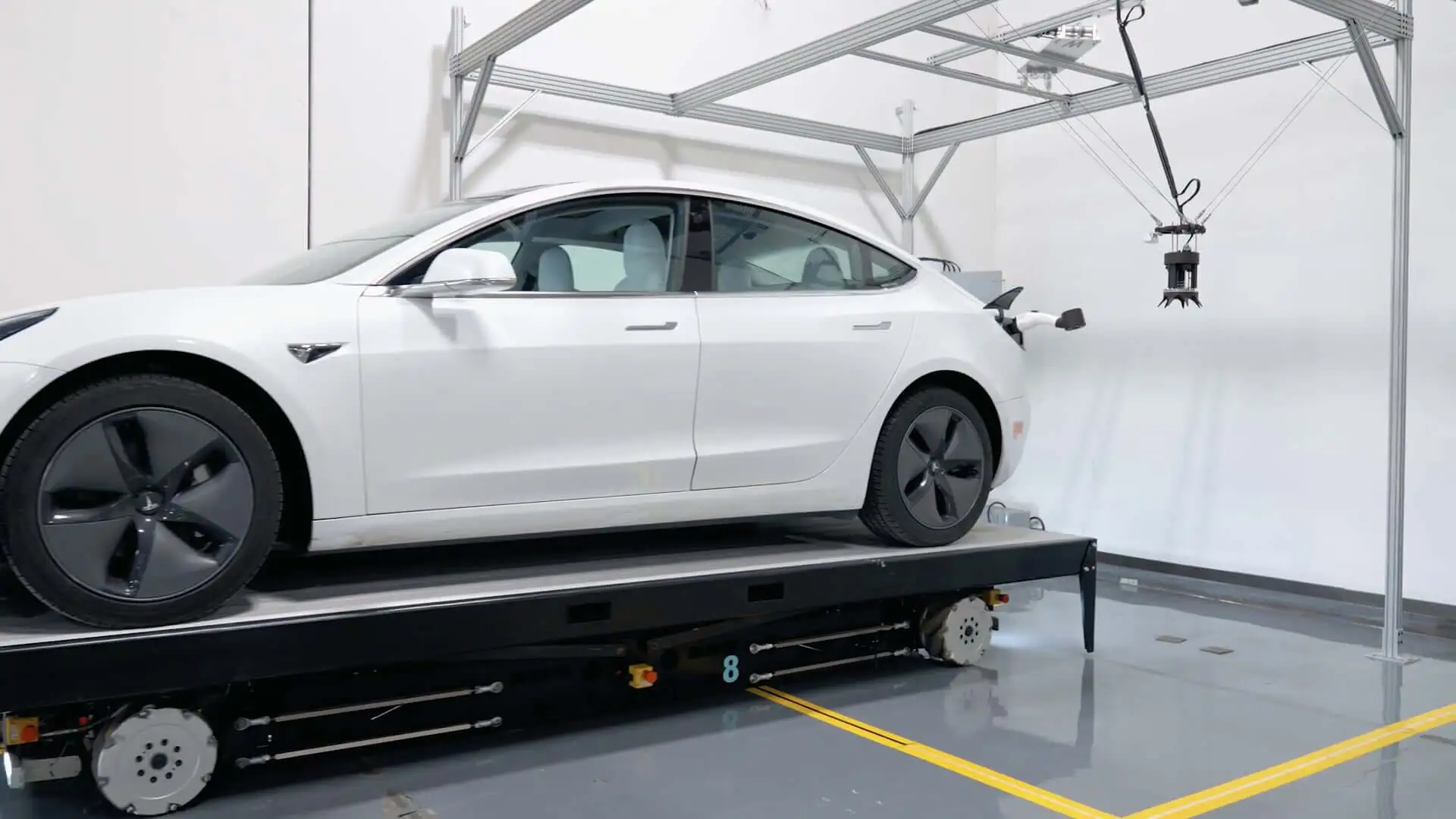 A white car parked on a metal platform inside a parking facility with automated EV charging equipment about to plug in for charging.
