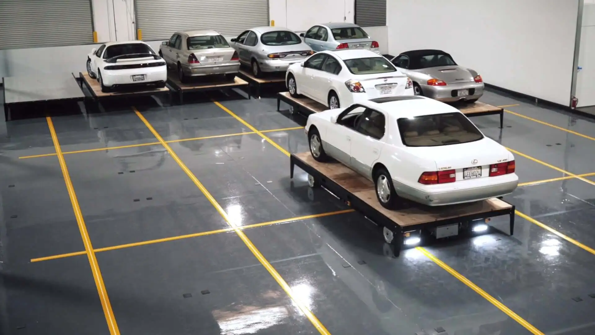 A white car mounted on a wooden platform with wheels in a spacious indoor parking lot, surrounded by other parked cars, with visible floor markings used by robots for navigation. A mobile robot is visible underneath the closest white car, driving the car to its parking spot