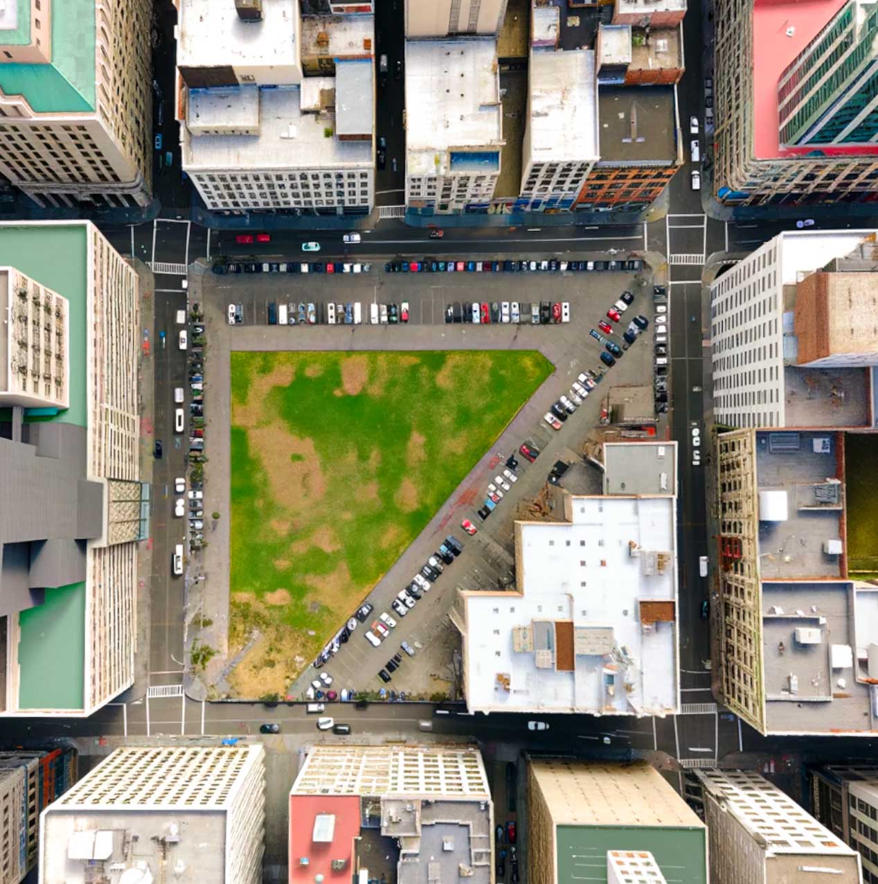 Aerial view of an urban city block with a green triangular park in the center, surrounded by tall buildings. Cars are parked along the perimeter of the park and on the surrounding streets. The buildings cast shadows, and some roads have visible crosswalks.
