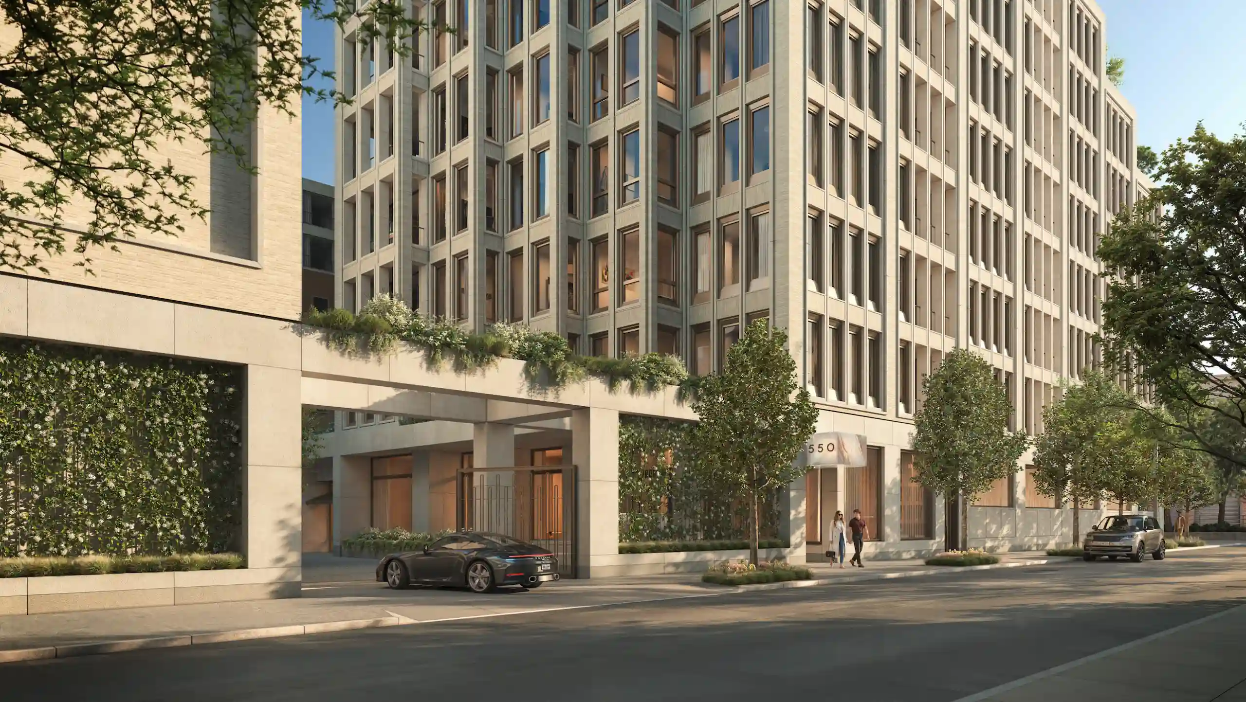 A modern multi-story building with large windows, greenery on ledges, trees along the sidewalk, a car parked on the street, and two people walking near the entrance.