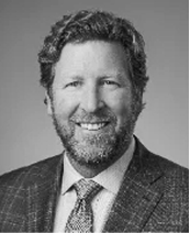 Black and white portrait of a man with curly hair and a beard, wearing a checked suit jacket, dress shirt, and patterned tie, smiling at the camera.