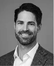 Black-and-white portrait of a smiling man with dark hair and a beard, wearing a textured suit jacket over a white shirt, posed in front of a plain background.