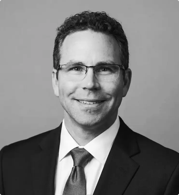 A man with short curly hair, glasses, and a suit jacket with a tie smiles at the camera against a plain background. The photo is black and white.