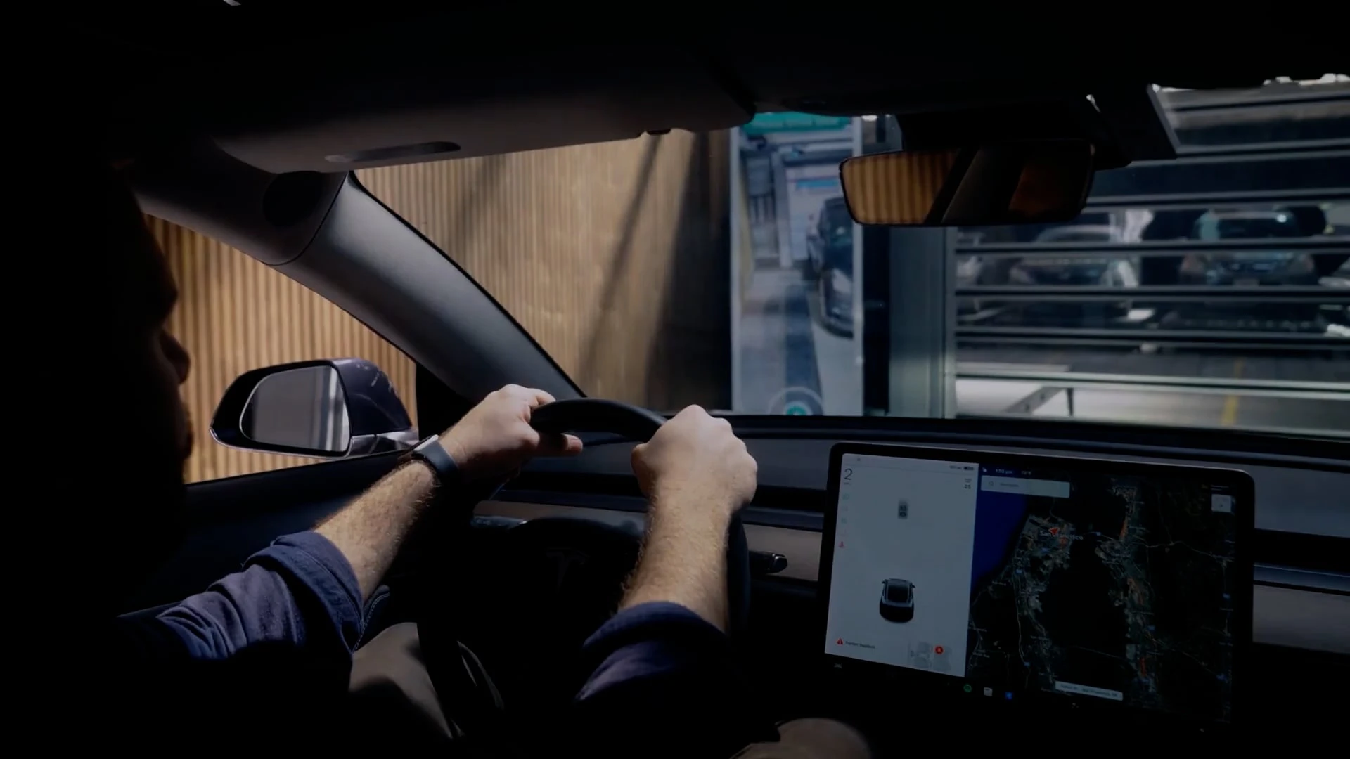 A person drives a Tesla car, viewed from the back seat, with hands on the steering wheel and a navigation map visible on the central touchscreen. As the car approaches a structure seen through the windshield, this scene exemplifies how automated parking systems work, seamlessly guiding vehicles toward designated spots with advanced technology.