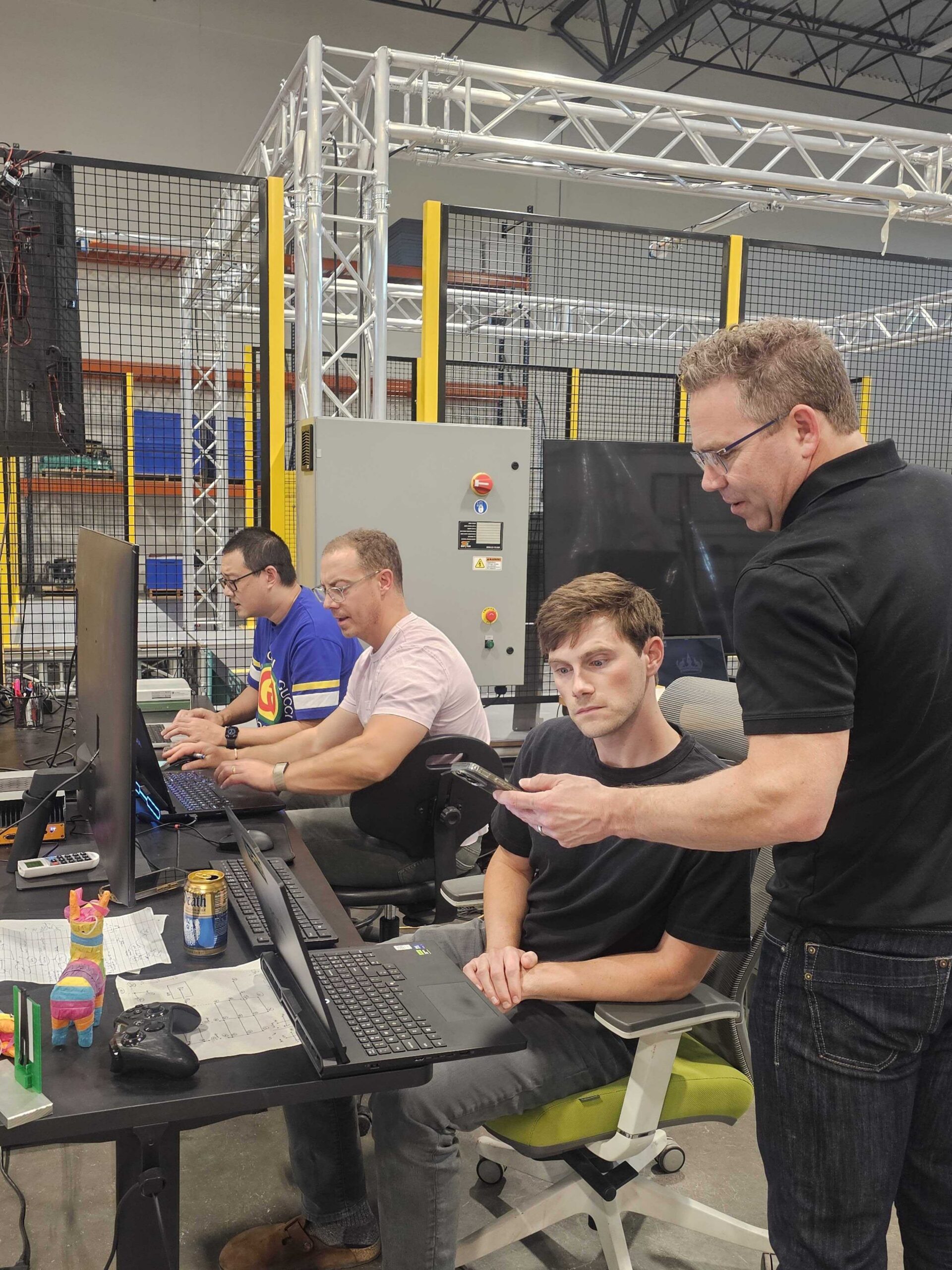 Four men work at computer stations in an industrial office, representing the dynamic environment of Careers at Volley Automation. Two type on keyboards, while one looks at a phone held by a standing colleague. Cables, monitors, and colorful desk toys are visible throughout the workspace.