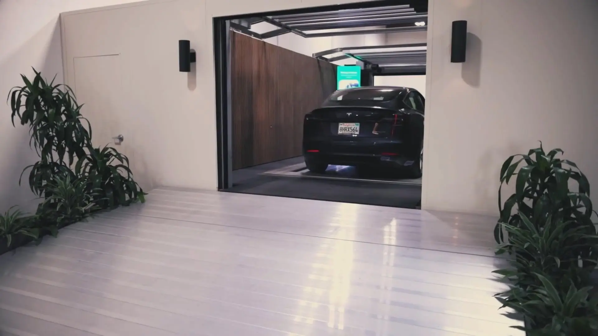 A black car is parked inside a modern garage with open doors, highlighting how automated parking systems work seamlessly in contemporary homes. Plants are placed on either side of the entrance, and the floor is clean and shiny.
