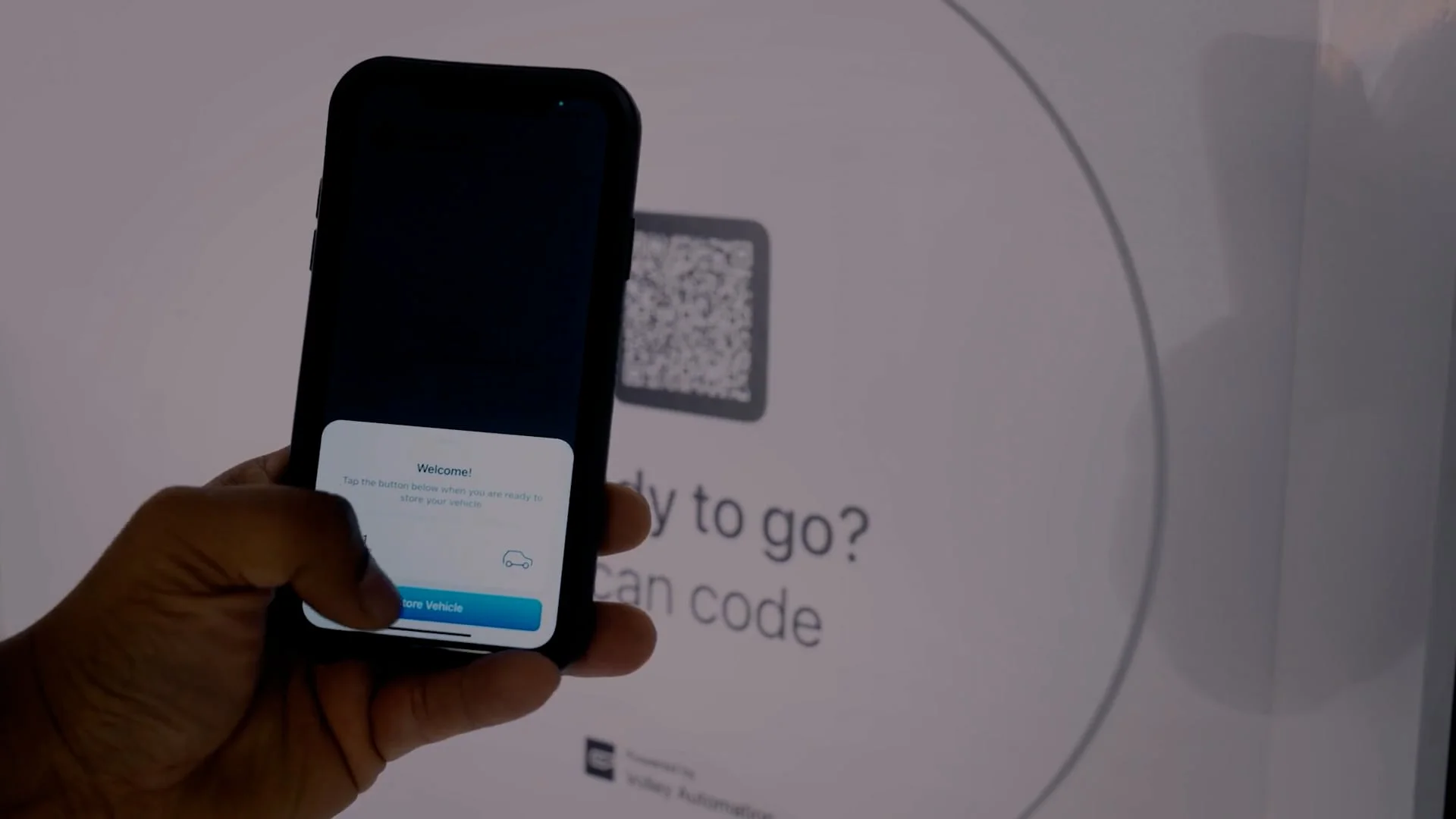 A hand holds a smartphone up to scan a QR code on a screen that reads “Ready to go? Scan code.” The phone displays a welcome message, illustrating how automated parking systems work seamlessly through digital access.