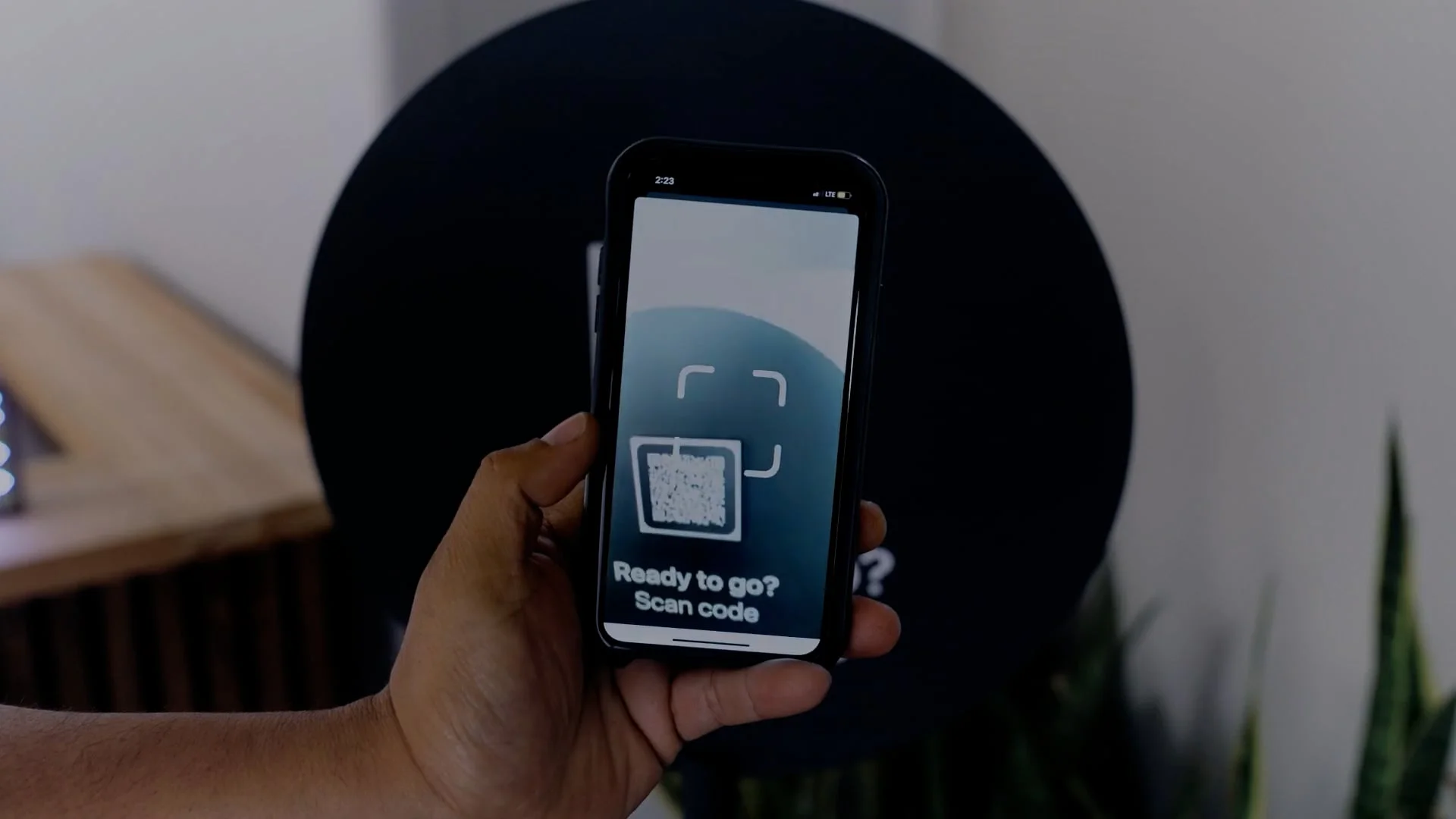 A hand holds a smartphone displaying a QR code scanner app with the text, “Ready to go? Scan code.” A dark circular object is blurred in the background, hinting at how automated parking systems work seamlessly with digital access.