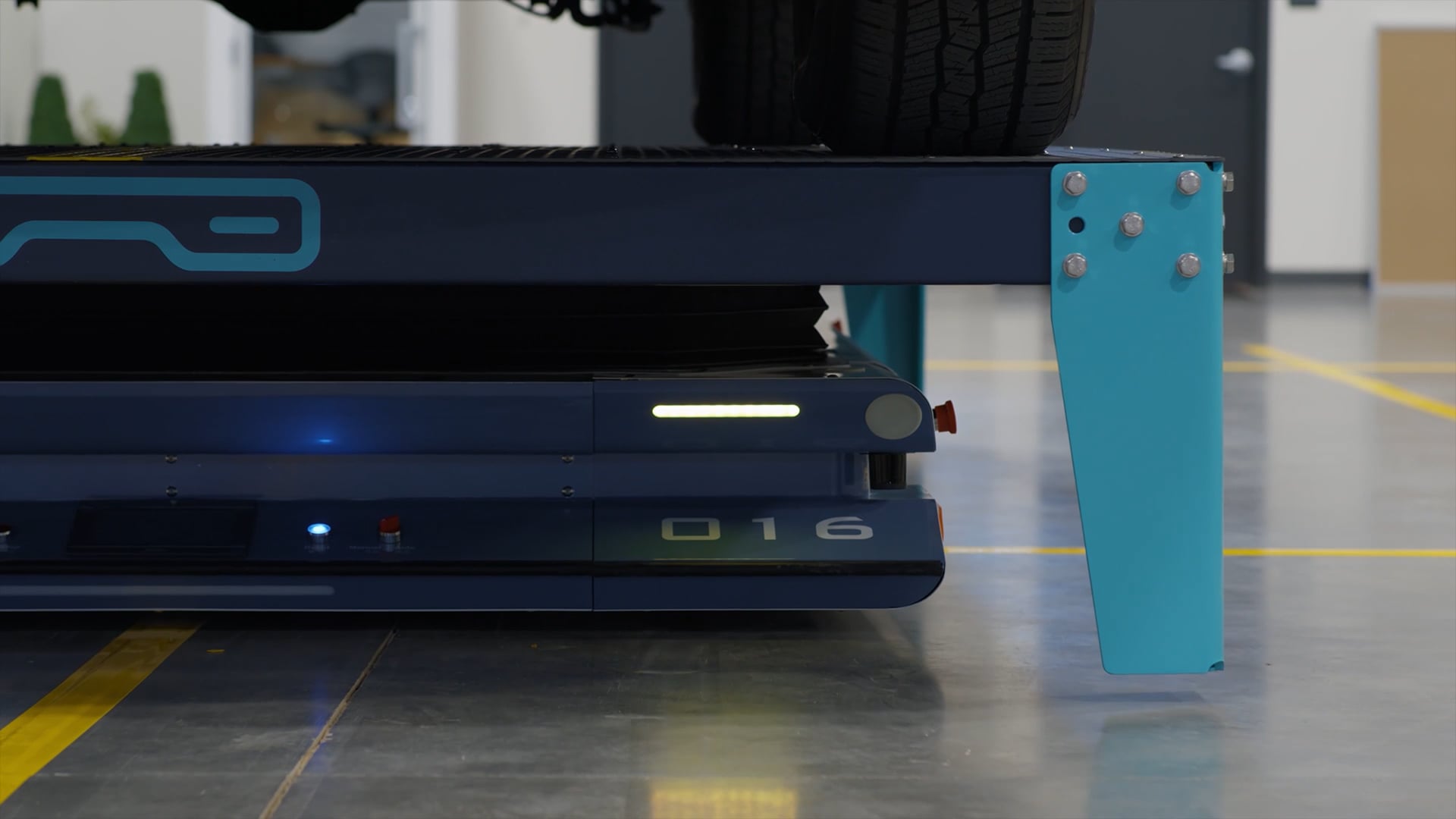 A close-up of a blue automated machine labeled “016” partially under a vehicle lift in a clean, modern garage with gray floors and blue structural supports, offering insight into how automated parking systems work.