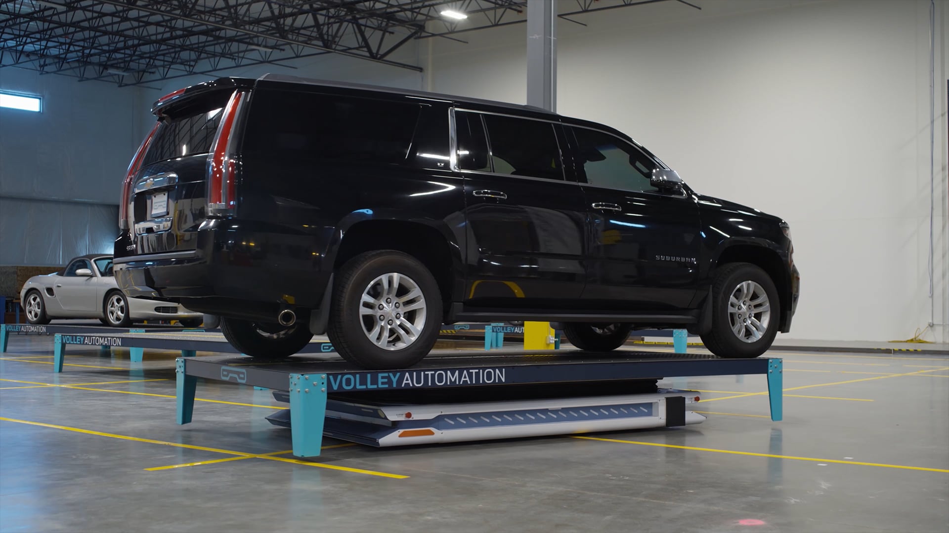 A black SUV is parked on a blue automated vehicle platform labeled “Volley Automation” inside a large, empty warehouse, illustrating how automated parking systems work to efficiently maneuver and store vehicles in shared spaces.