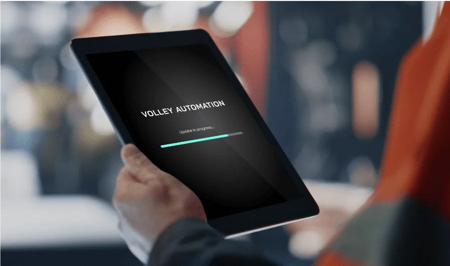 A person holds a tablet displaying a screen with the words “VOLLEY AUTOMATION” and a loading bar below labeled “Update in progress,” highlighting the process of working with Volley Automation. The background is blurred.