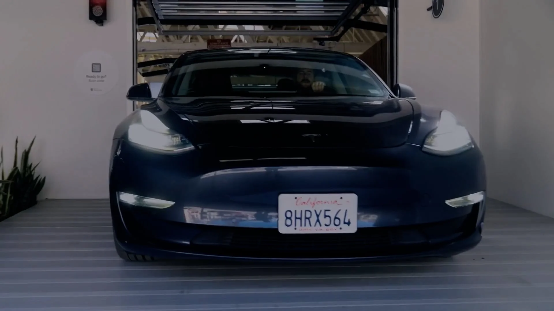 A black Tesla car with headlights on is parked in a garage, illustrating how automated parking systems work in modern vehicles. California license plate 8HRX564 is visible on the front. Some plants are on the left side of the garage.