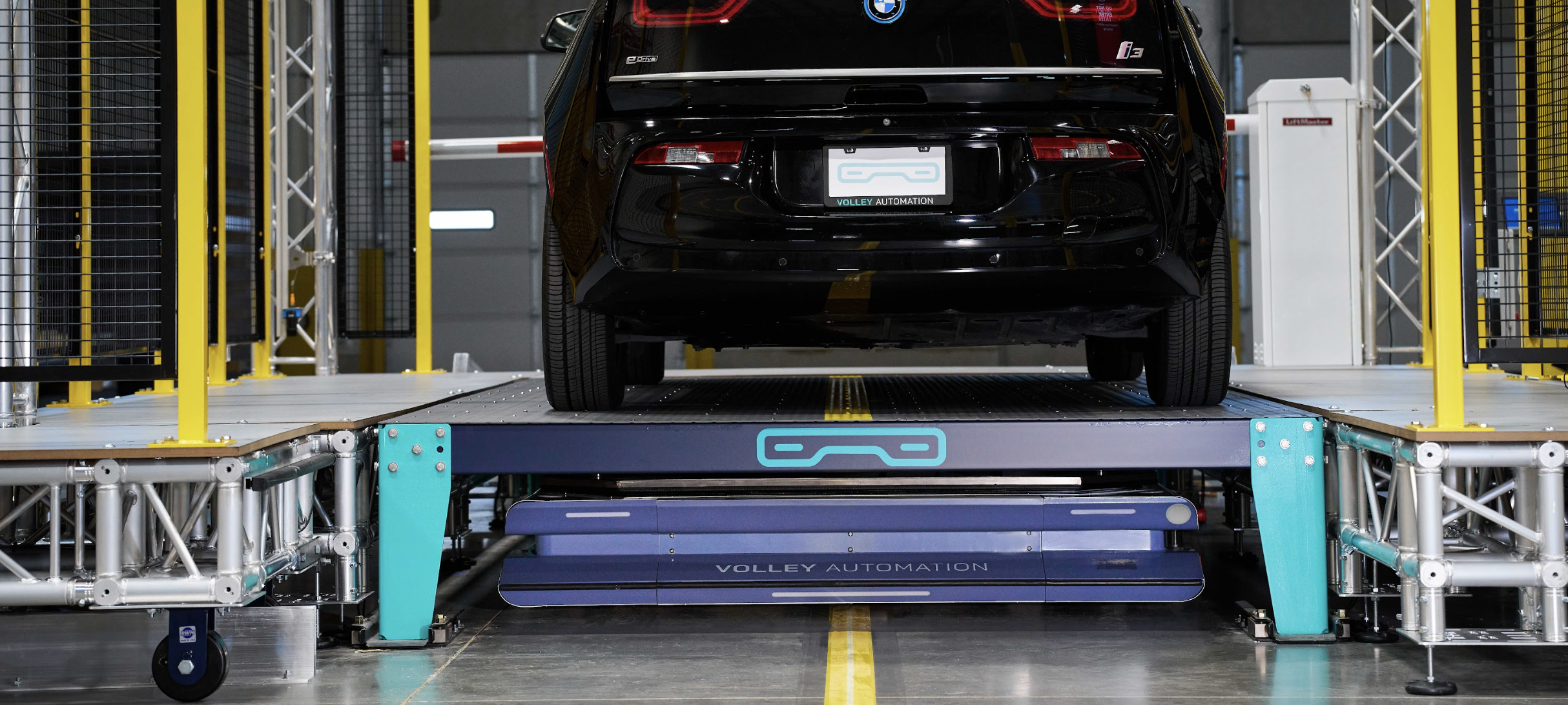 A black car is parked on a metal platform labeled “Volley Automation,” inside a modern industrial facility with yellow railings and safety barriers, showcasing the precision and innovative solutions that come from working with Volley Automation.