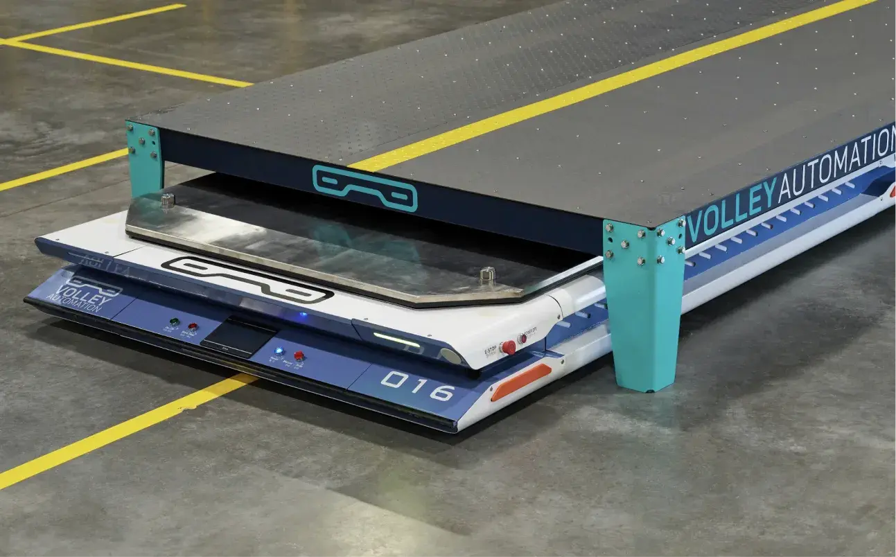 A compact, flat autonomous robot labeled “016” moves under a metal platform in a warehouse setting, demonstrating principles similar to how automated parking systems work—efficiently handling and transporting materials without human intervention.