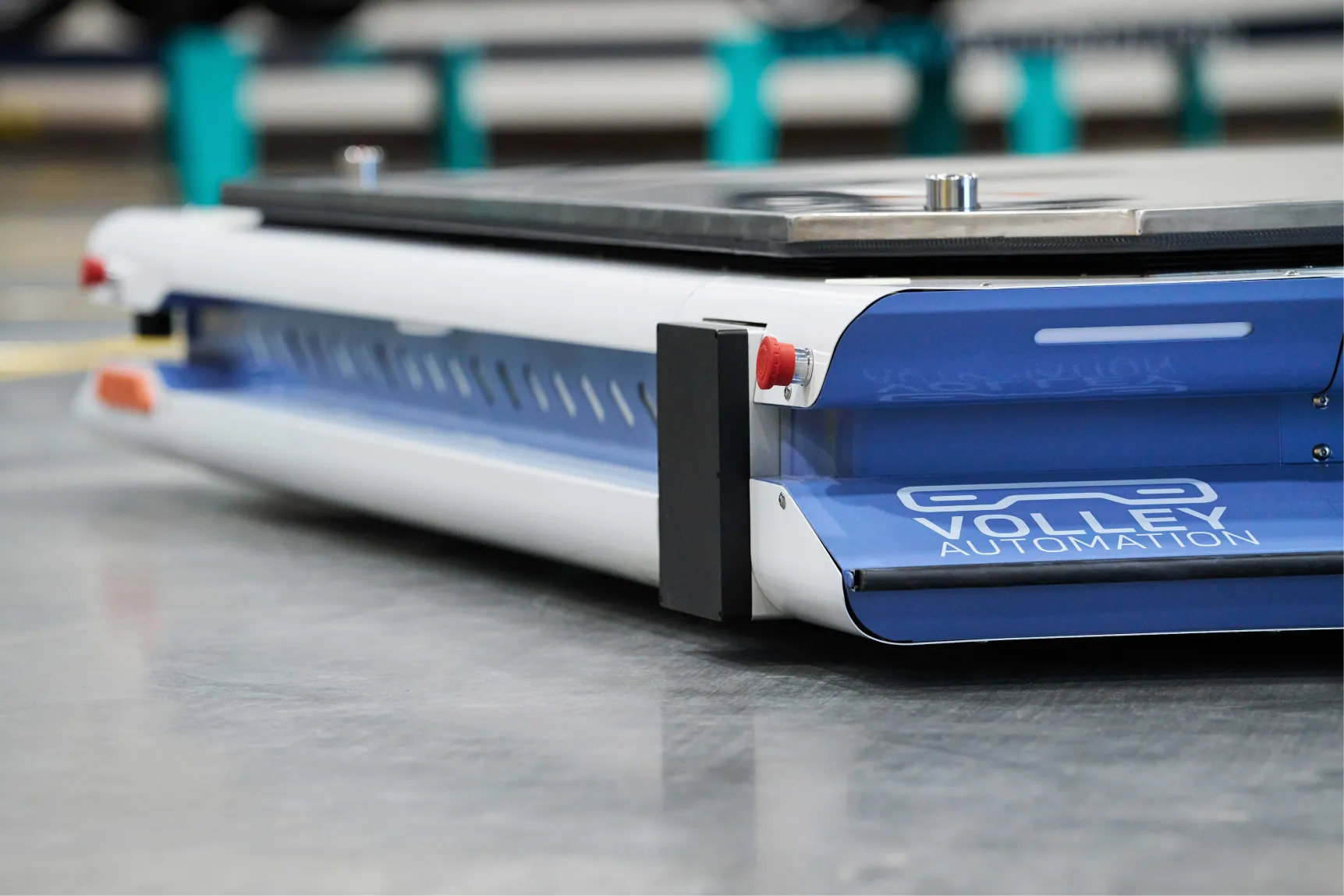A close-up of a blue and white automated guided vehicle (AGV) labeled “Volley Automation” on a smooth industrial floor, with blurred equipment in the background, showcases the advanced technology behind modern logistics—similar to how automated parking systems work to efficiently transport vehicles within tight spaces.