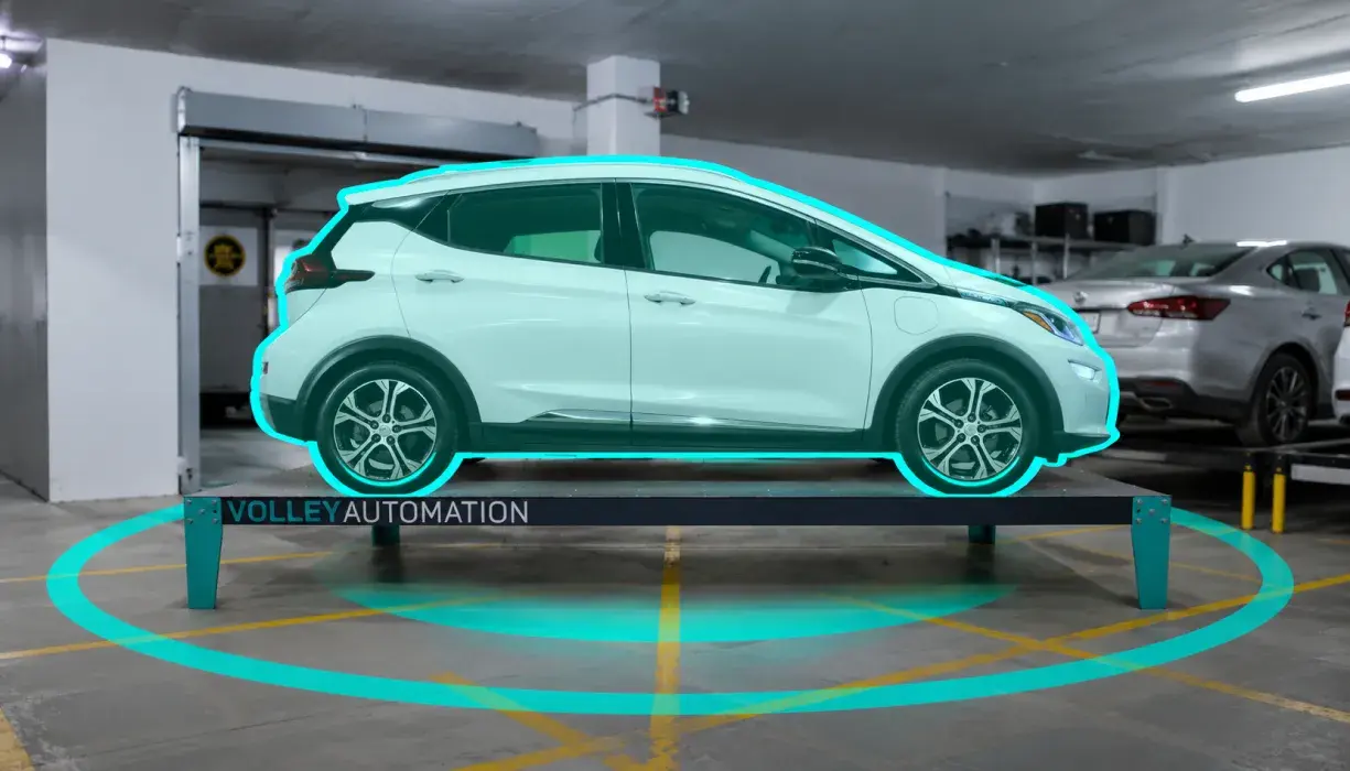 A white electric car outlined in blue is parked on a metal platform inside a parking garage, with a blue circle projected on the floor around it, offering an impressive glimpse into how automated parking systems work.